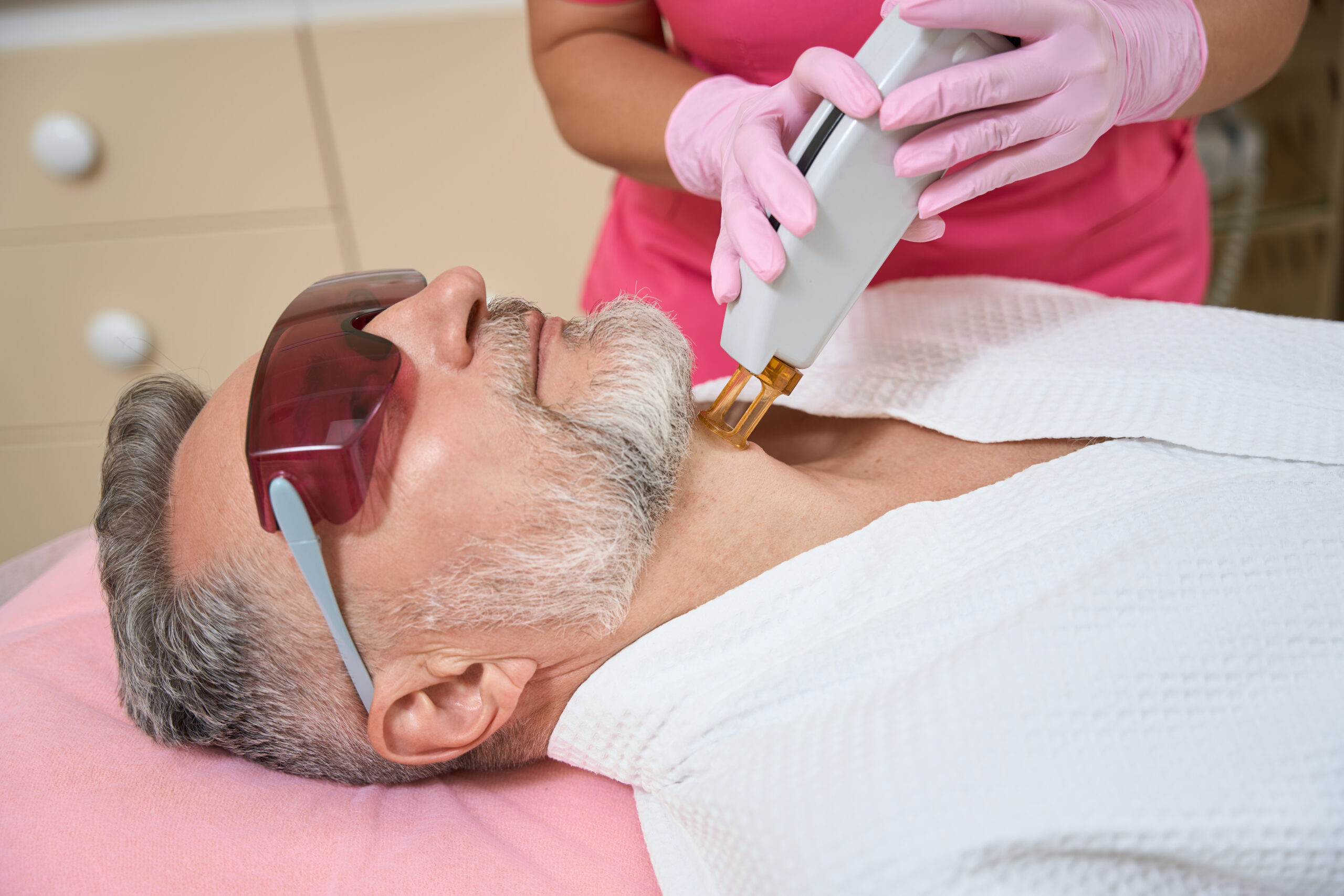 Female hands of cosmetologist doing laser epilation to bearded man on his neck in beauty salon. Cropped photo
