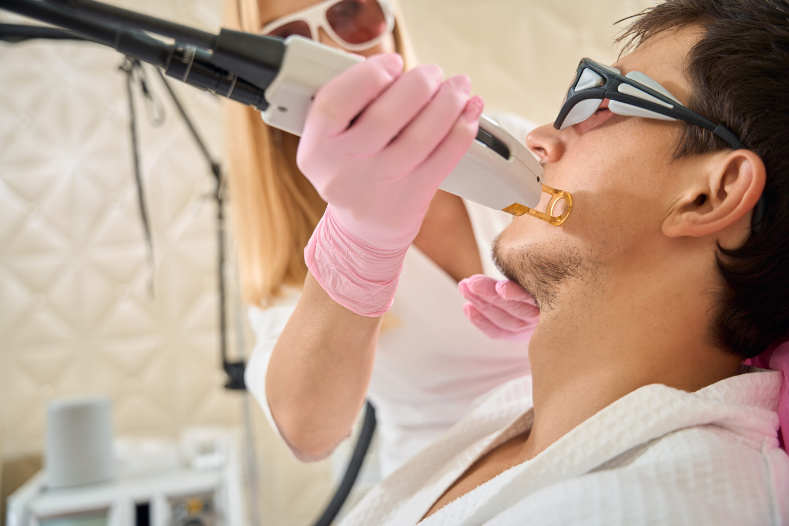 Female performs facial laser hair removal procedure on young client, people wearing safety glasses